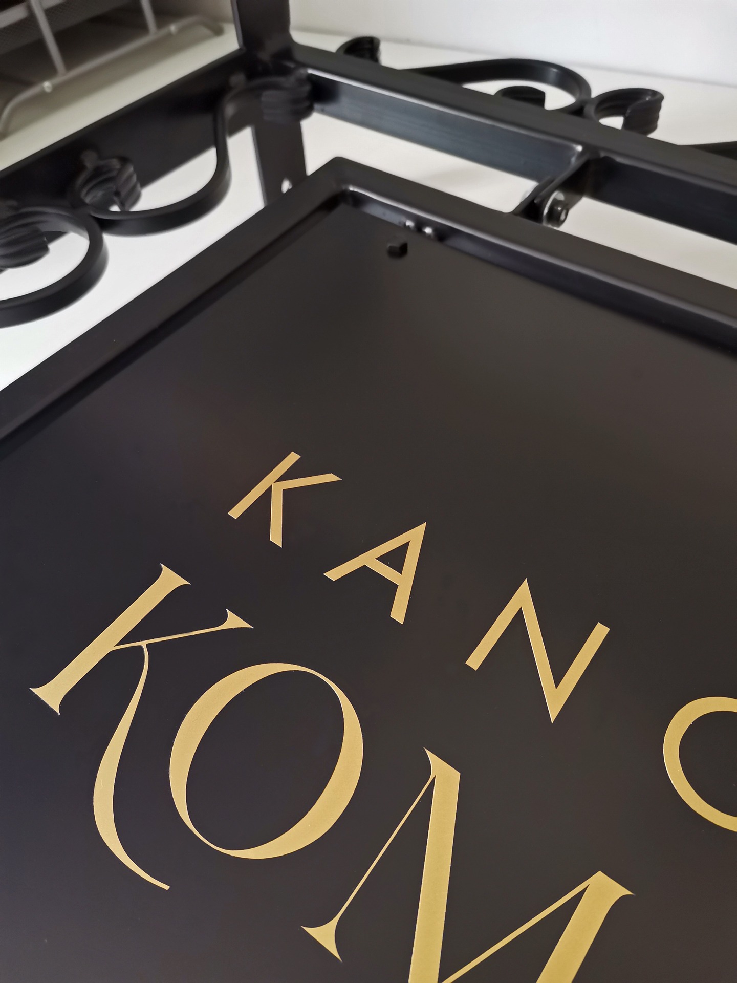 Czarny szyld z napisem 'KANCELARIA' wykonanym złotą czcionką, zawieszony na ozdobnym, kutym uchwycie.