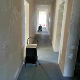 Dwa przemysłowe osuszacze powietrza ustawione w surowym, długim korytarzu w trakcie remontu. Ściany z widocznymi śladami wilgoci i niedokończonymi pracami wykończeniowymi.