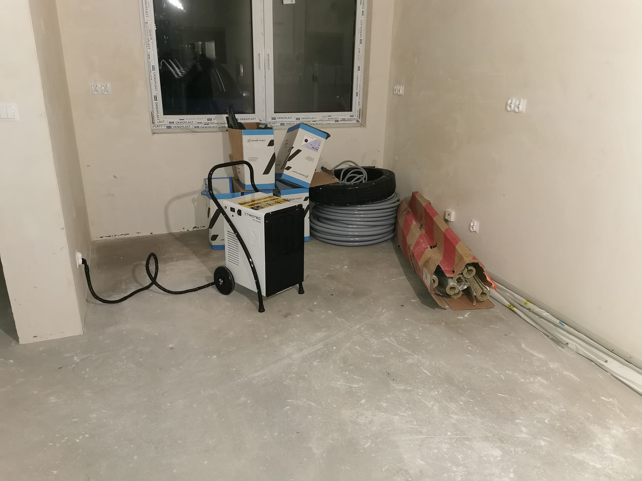Przenośny osuszacz powietrza Trotec w surowym pomieszczeniu z widocznymi śladami wilgoci na ścianach i podłodze, obok kartony i rury instalacyjne.