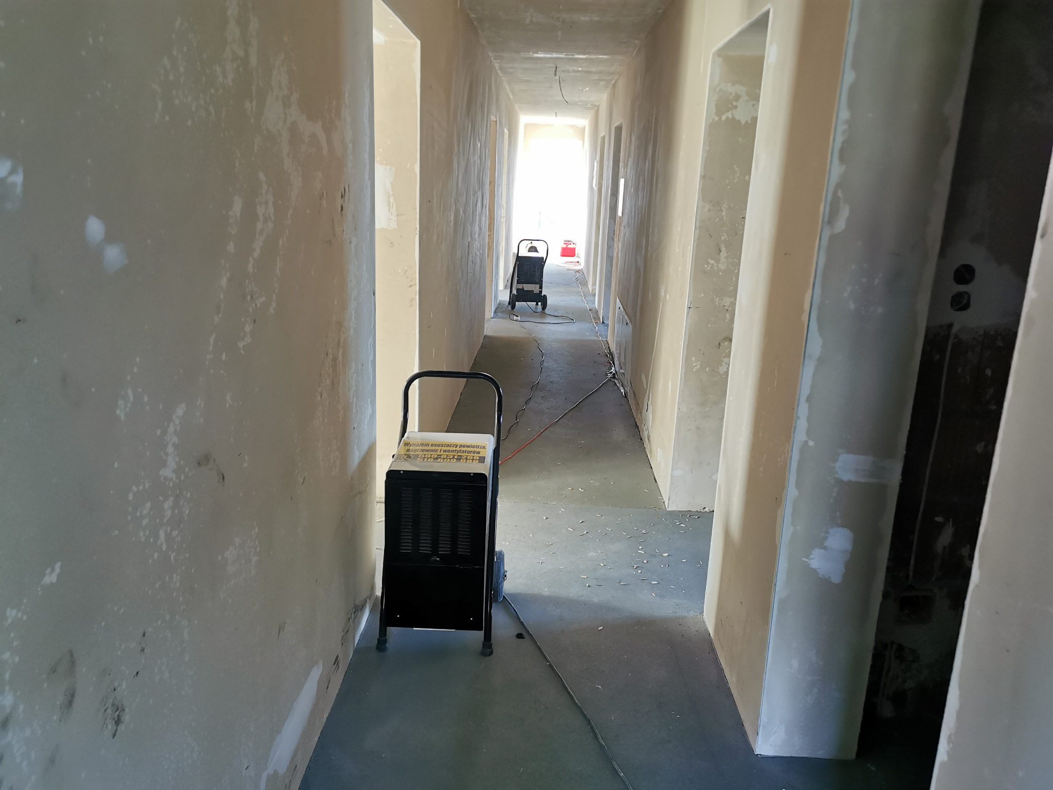 Dwa przemysłowe osuszacze powietrza ustawione w surowym, długim korytarzu w trakcie remontu. Ściany z widocznymi śladami wilgoci i niedokończonymi pracami wykończeniowymi.
