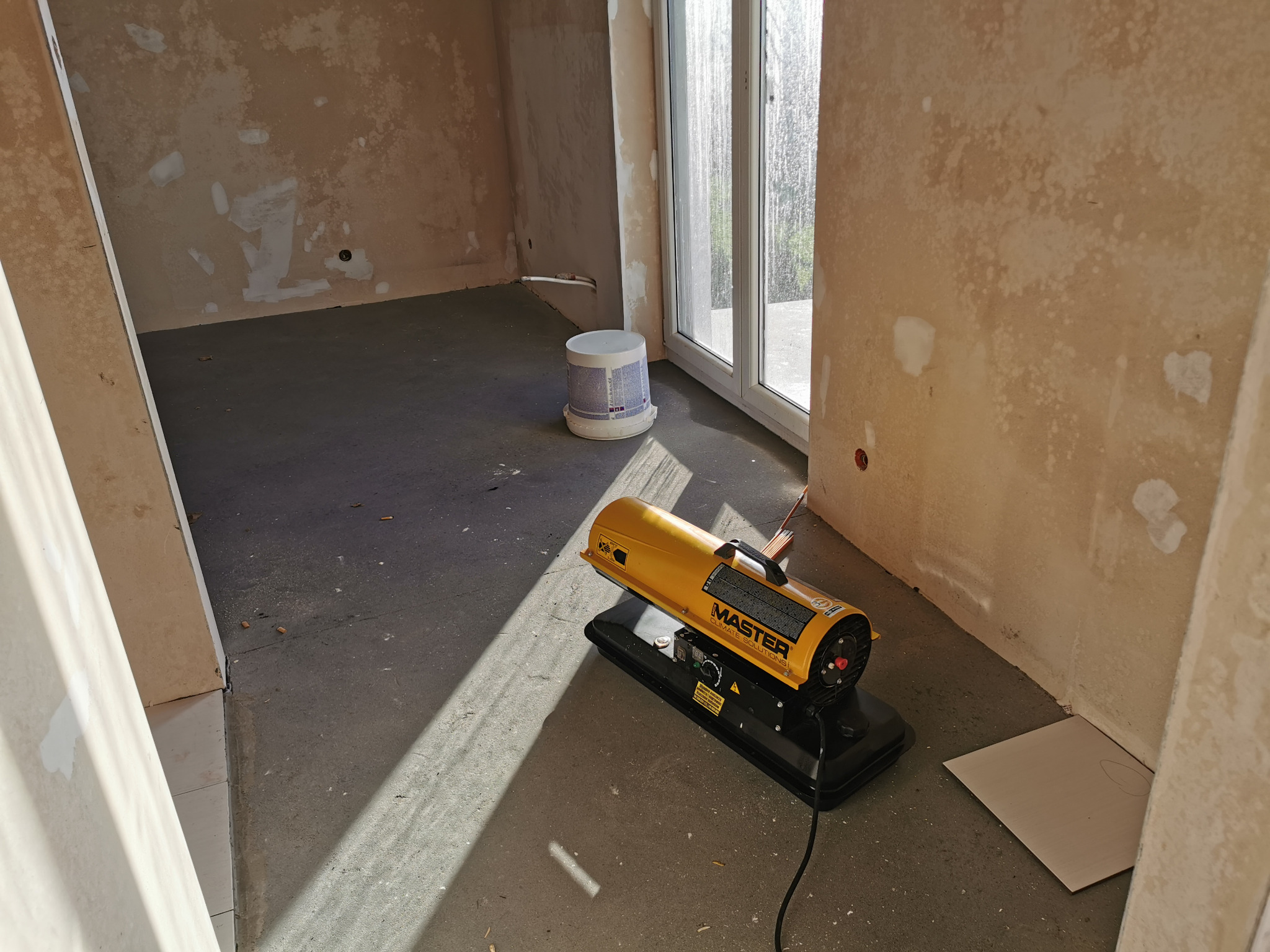 Żółta nagrzewnica MASTER na czarnej podstawie, ustawiona w surowym pomieszczeniu z oknem i wiadrem, ślady po naprawach na ścianach.