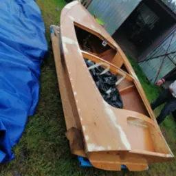 Drewniana łódź w trakcie renowacji, widoczne szpachlowanie i przygotowanie do malowania, na trawie obok leży niebieska plandeka.