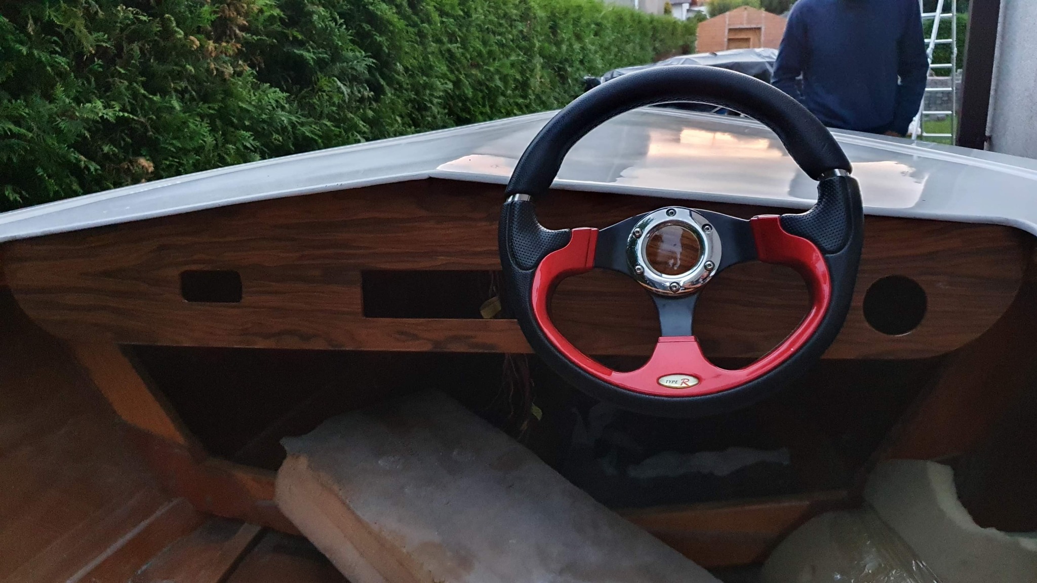 Wnętrze łodzi motorowej z drewnianą deską rozdzielczą i sportową kierownicą z czerwonymi akcentami, widoczne fragmenty laminatu, w tle zieleń.