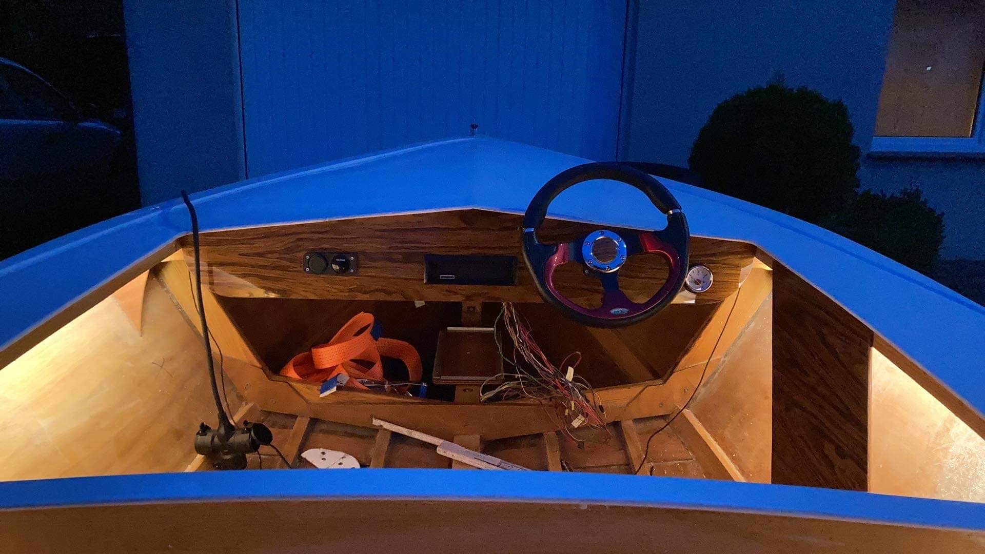 Wnętrze łodzi w trakcie renowacji, widoczne drewniane elementy konstrukcyjne, panel sterowania z kierownicą i wskaźnikami, oraz luźno zwisające przewody elektryczne. Pomarańczowy pas transportowy...