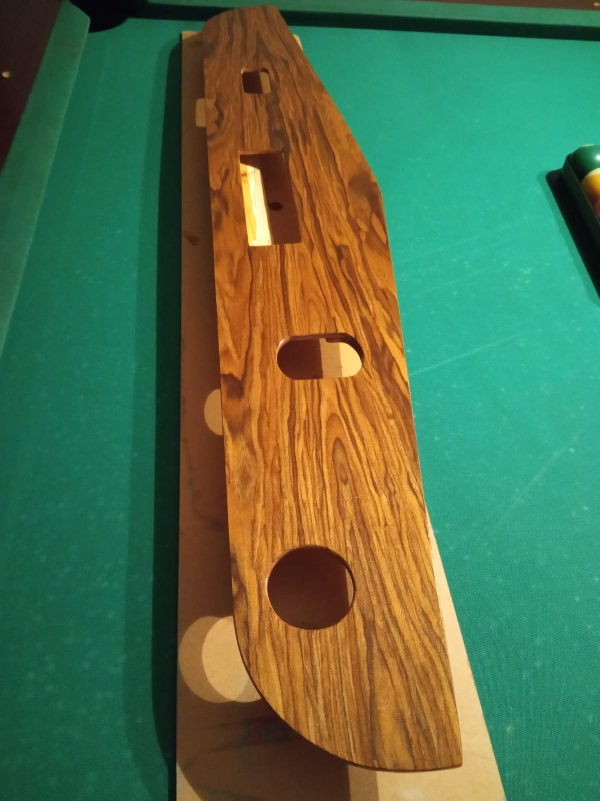 Drewniana forma z otworami i wycięciami, leżąca na zielonym stole bilardowym, ukazująca strukturę drewna i proces obróbki