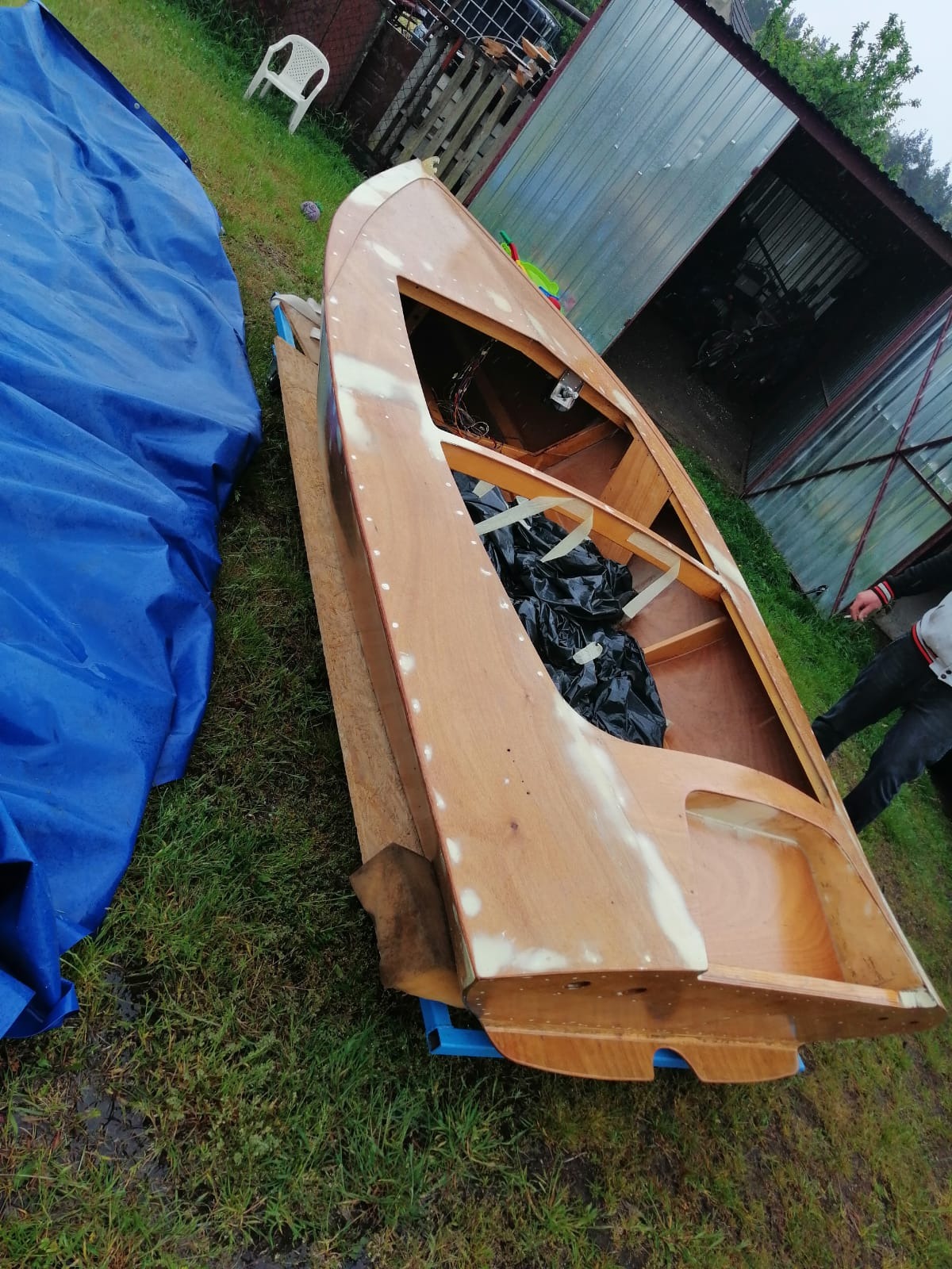 Drewniana łódź w trakcie renowacji, widoczne szpachlowanie i przygotowanie do malowania, na trawie obok leży niebieska plandeka.