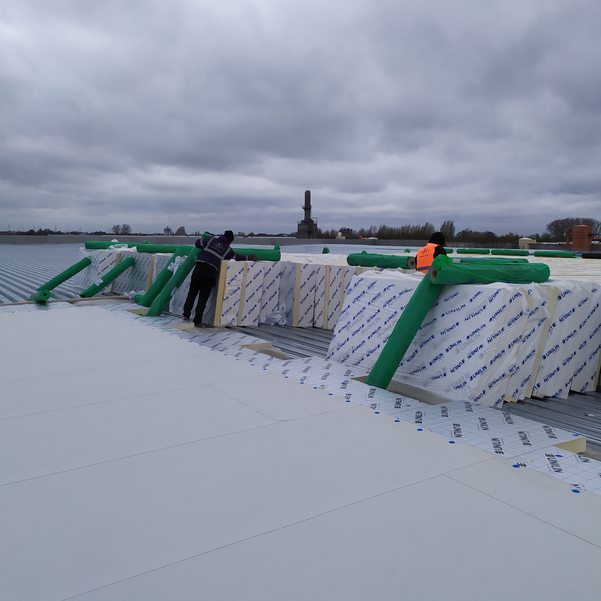 Montaż izolacji termicznej z płyt styropianowych na dachu przemysłowym, widoczni pracownicy w odzieży roboczej, przygotowane rolki folii ochronnej i komin w tle.