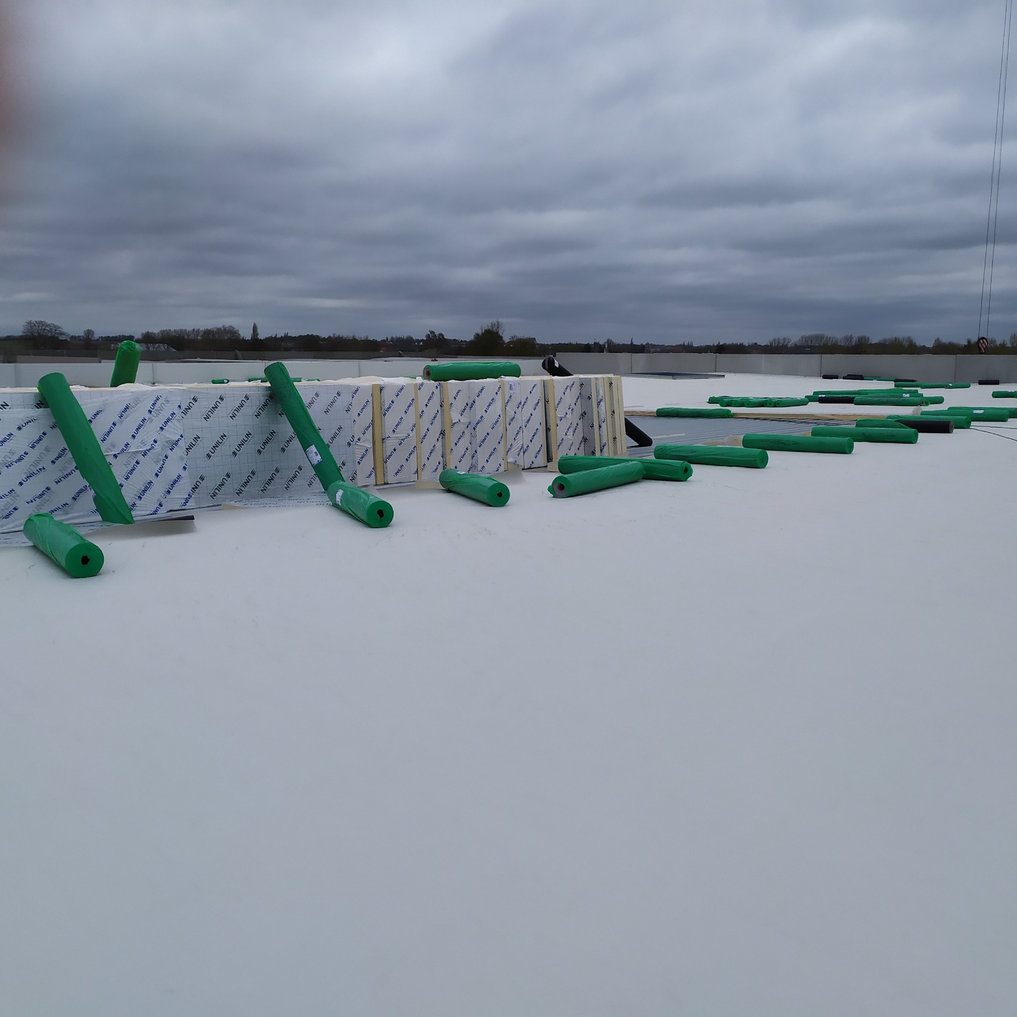 Biała membrana dachowa z rozłożonymi rolkami zielonej folii ochronnej i częściowo ułożonymi płytami izolacyjnymi z widocznym nadrukiem producenta, ujęcie z góry przy pochmurnym niebie.