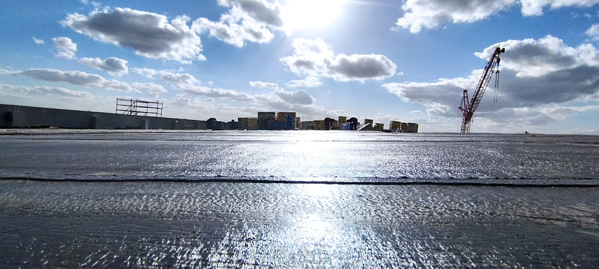 Błyszcząca, świeżo położona izolacja na dachu budynku w słoneczny dzień, na horyzoncie widoczne budynki i żuraw budowlany, błękitne niebo z chmurami.