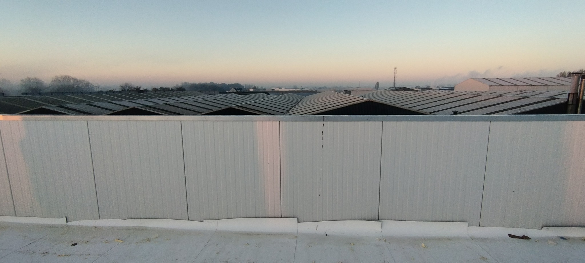 Widok z góry na przemysłowe dachy pokryte blachą trapezową, z lekką szadzią, w tle zamglone drzewa i budynki, na pierwszym planie biała ściana z blachy falistej.