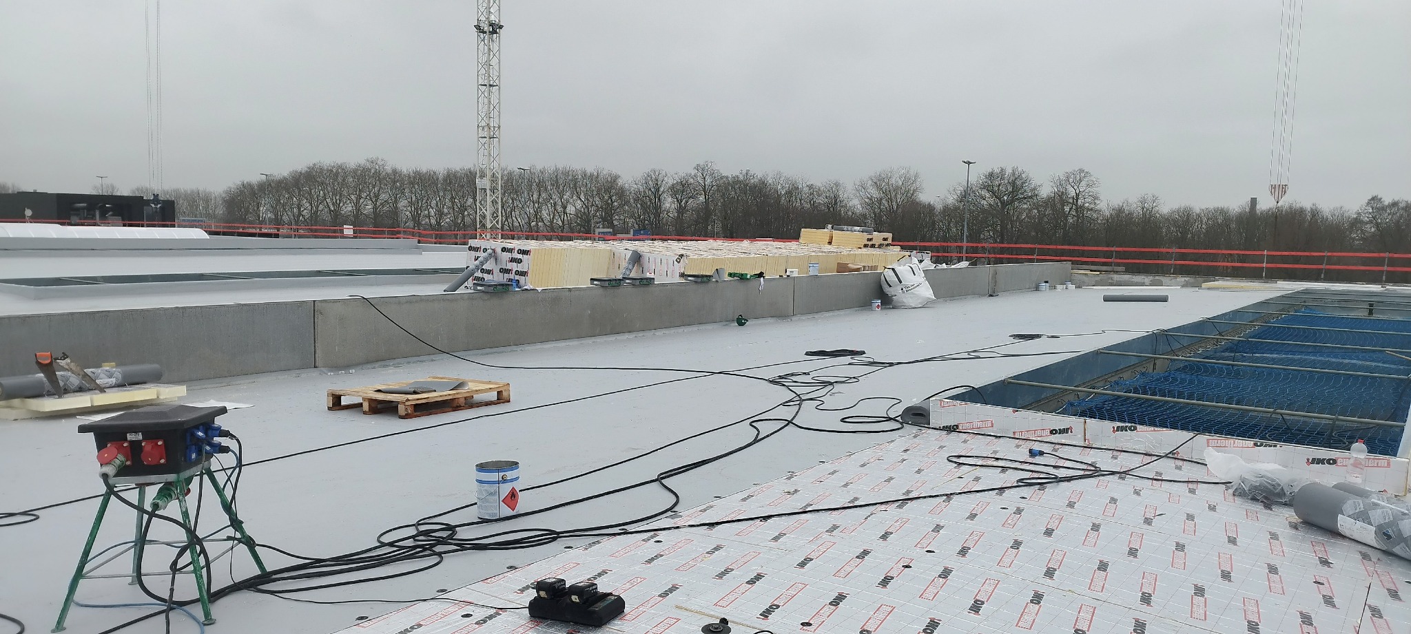 Rozległy płaski dach w trakcie hydroizolacji, widoczne rolki papy termozgrzewalnej z nadrukiem, rozdzielnica elektryczna na trójnogu, paleta, puszka z klejem, w oddali przygotowane płyty izolacyjne...