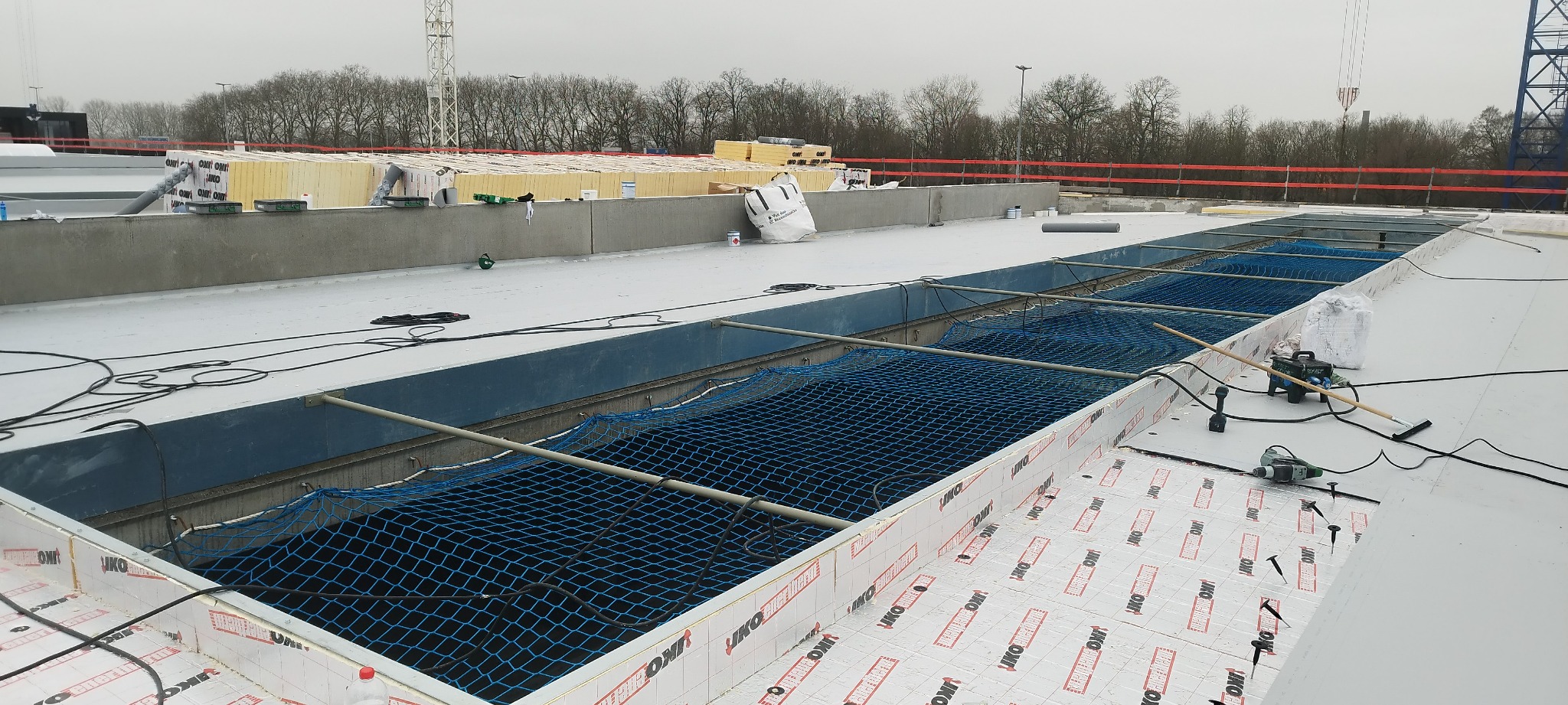 Montaż zabezpieczenia siatką asekuracyjną w otworze na dachu płaskim, widoczne narzędzia i materiały izolacyjne