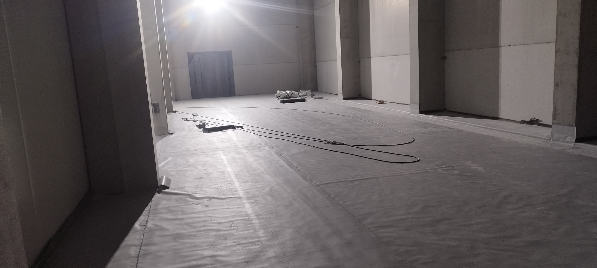 Wnętrze hali produkcyjnej z betonową posadzką pokrytą szarą izolacją przeciwwilgociową, widoczne kable i rolka materiału izolacyjnego w tle.