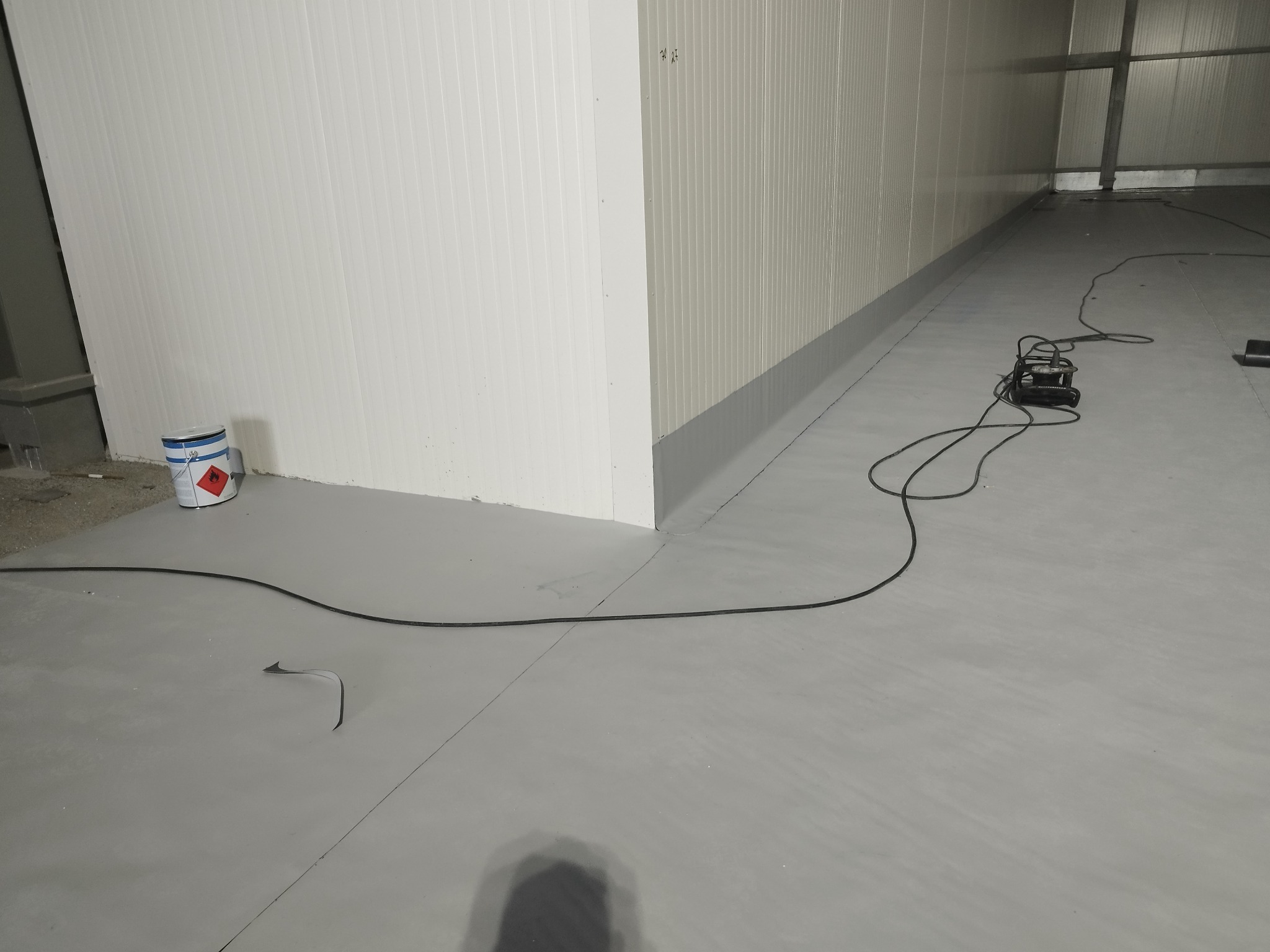 Wnętrze hali przemysłowej z białymi ścianami z paneli i szarą, świeżo wylaną posadzką żywiczną; widoczny agregat szlifierski z kablem oraz puszka farby przy ścianie.