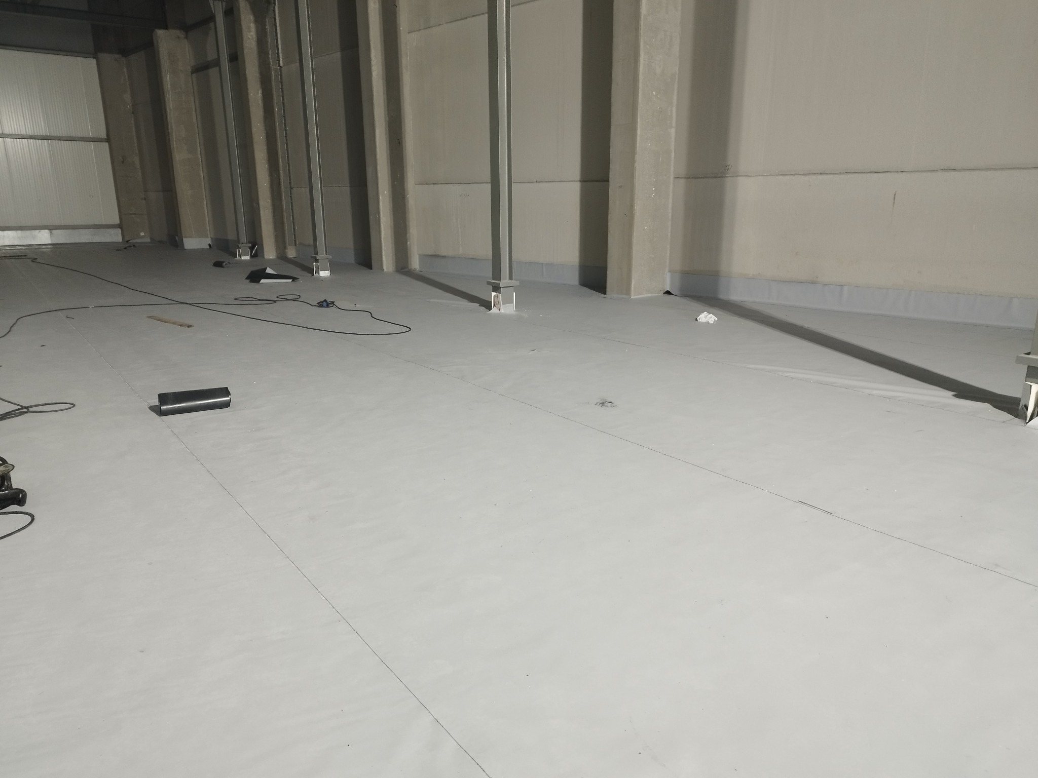 Wnętrze hali przemysłowej z szarą, gładką posadzką, widoczne metalowe słupy konstrukcyjne i narzędzia na podłodze.