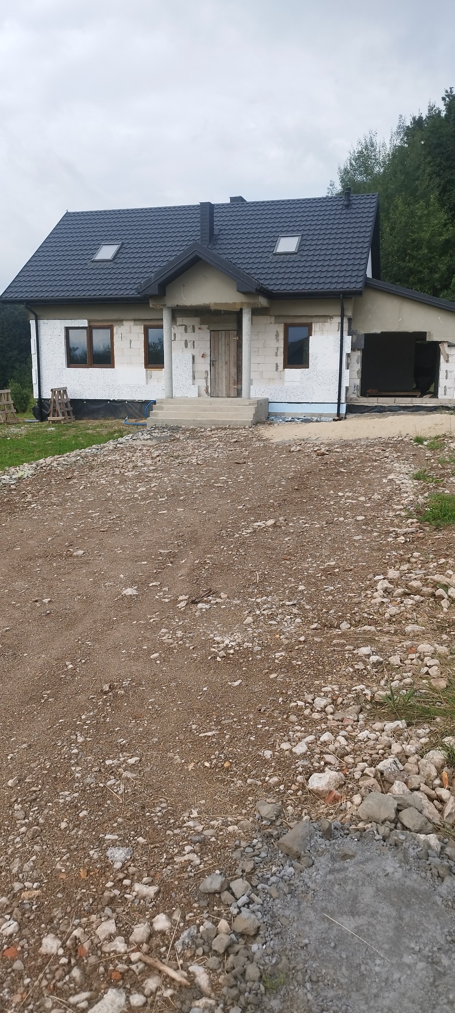 Dom jednorodzinny w trakcie ocieplania elewacji, widoczne płyty izolacyjne na ścianach i nowy dach z oknami dachowymi, teren wokół domu częściowo utwardzony.