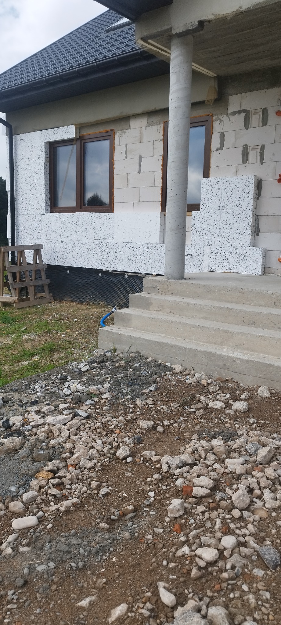 Niedokończona elewacja budynku z widocznym styropianem, częściowo ocieplona ściana, betonowe schody wejściowe i gruz na ziemi.