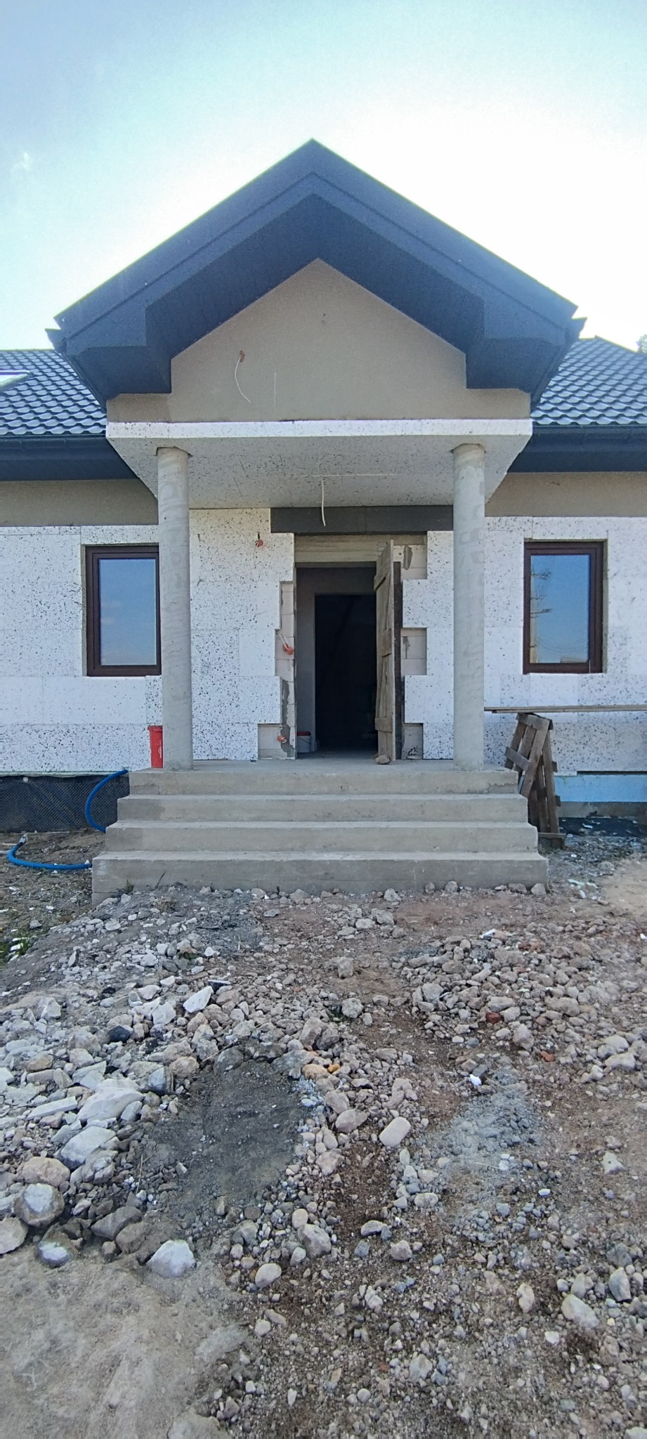 Dom jednorodzinny w trakcie ocieplania elewacji, widoczne białe płyty izolacyjne, betonowe kolumny i schody wejściowe, teren wokół budynku z gruzem i kamieniami.