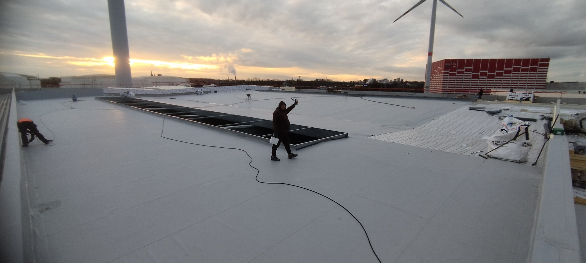 Ekipa dekarzy na rozległym, płaskim dachu pokrytym szarą membraną, z widocznymi elementami konstrukcyjnymi i wentylacyjnymi, turbiny wiatrowe i budynki w tle, wieczorne światło.