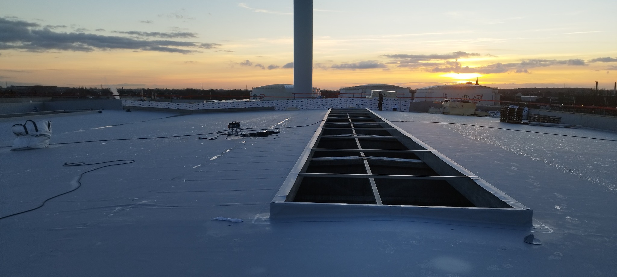Biała membrana dachowa z widocznym szkieletem świetlika i zgrzewarką w tle, na tle zachodzącego słońca i zbiorników przemysłowych.