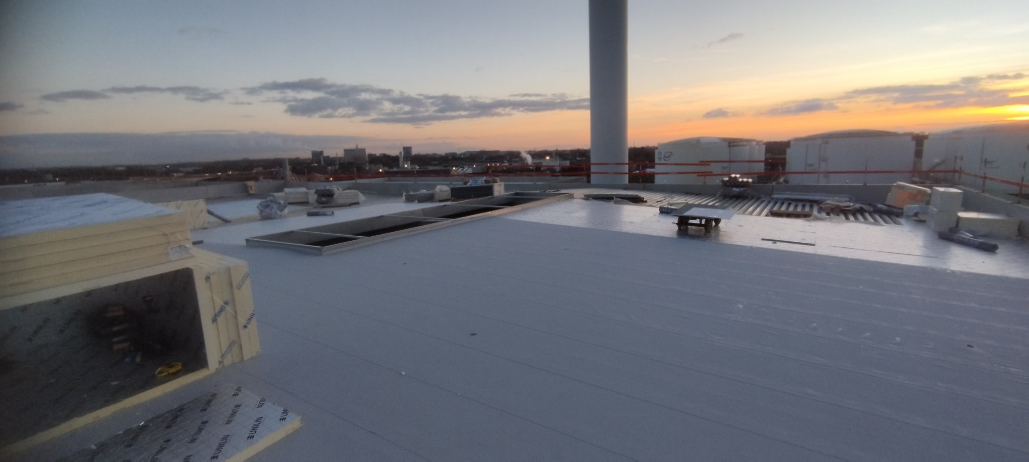Widok z góry na płaski dach pokryty szarą membraną, z widocznymi świetlikami i elementami wentylacji; w tle zachód słońca nad miastem.