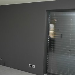 SKYLINE MONKEY ELGAS PIOTR - Świeżo pomalowane na szaro wnętrze z widocznymi gniazdkami, fragmentem podłogi z płytkami i oknem z żaluzjami, surowa żarówka zwisa z sufitu.