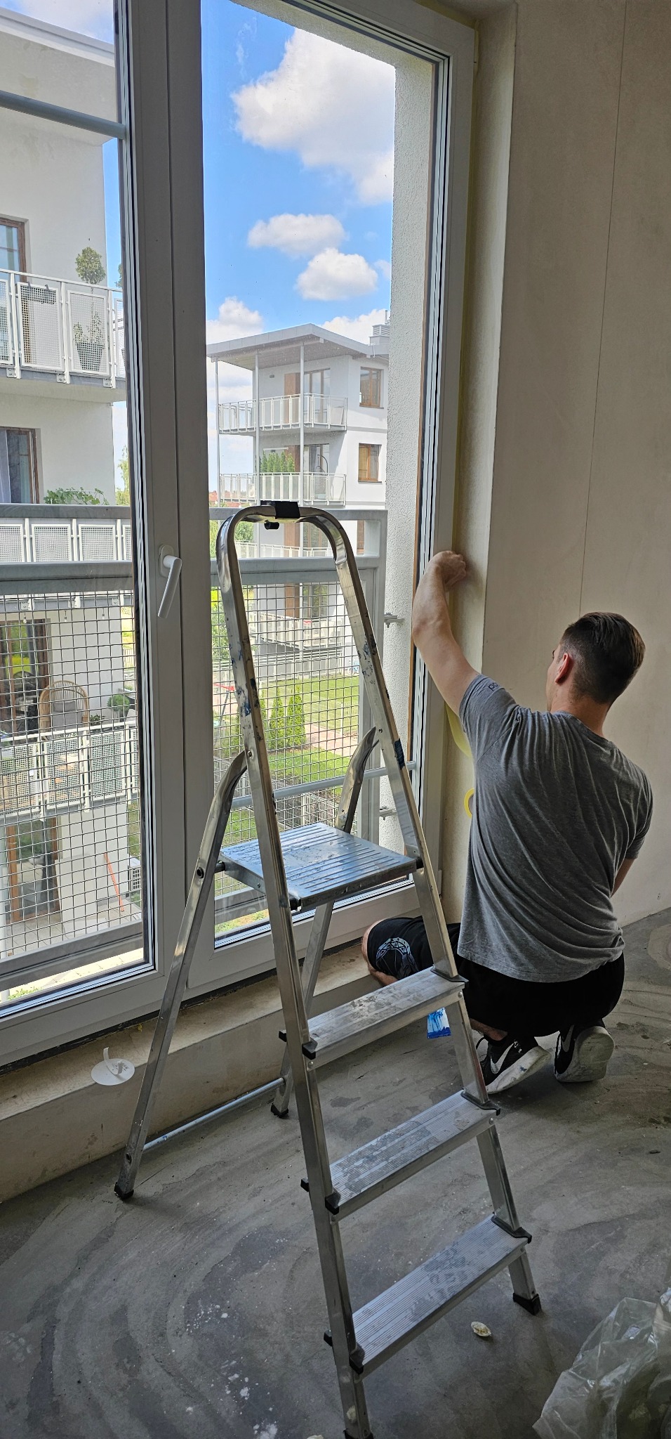 Pracownik na kolanach przy oknie, zabezpiecza folią malarską ramę okienną przed malowaniem, widoczna aluminiowa drabina i blok mieszkalny za oknem.