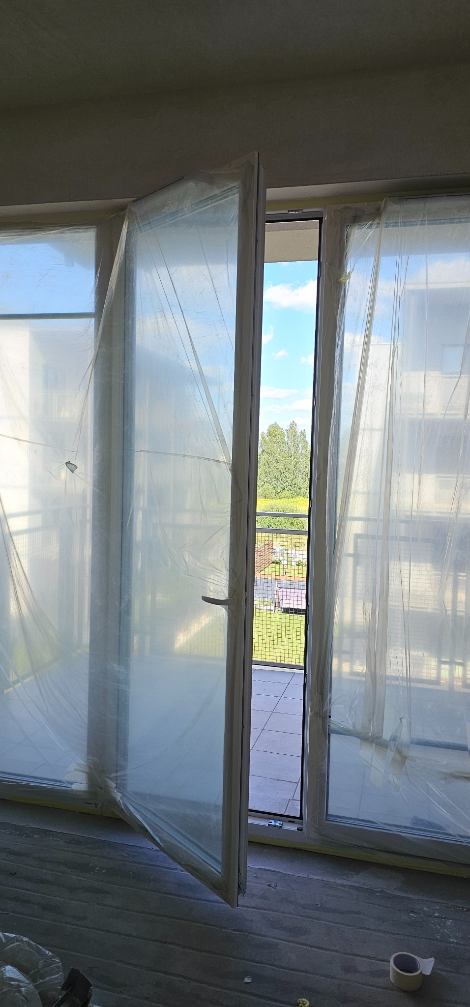 Okna balkonowe zabezpieczone folią malarską przed remontem, widok na balkon i zieleń na zewnątrz.