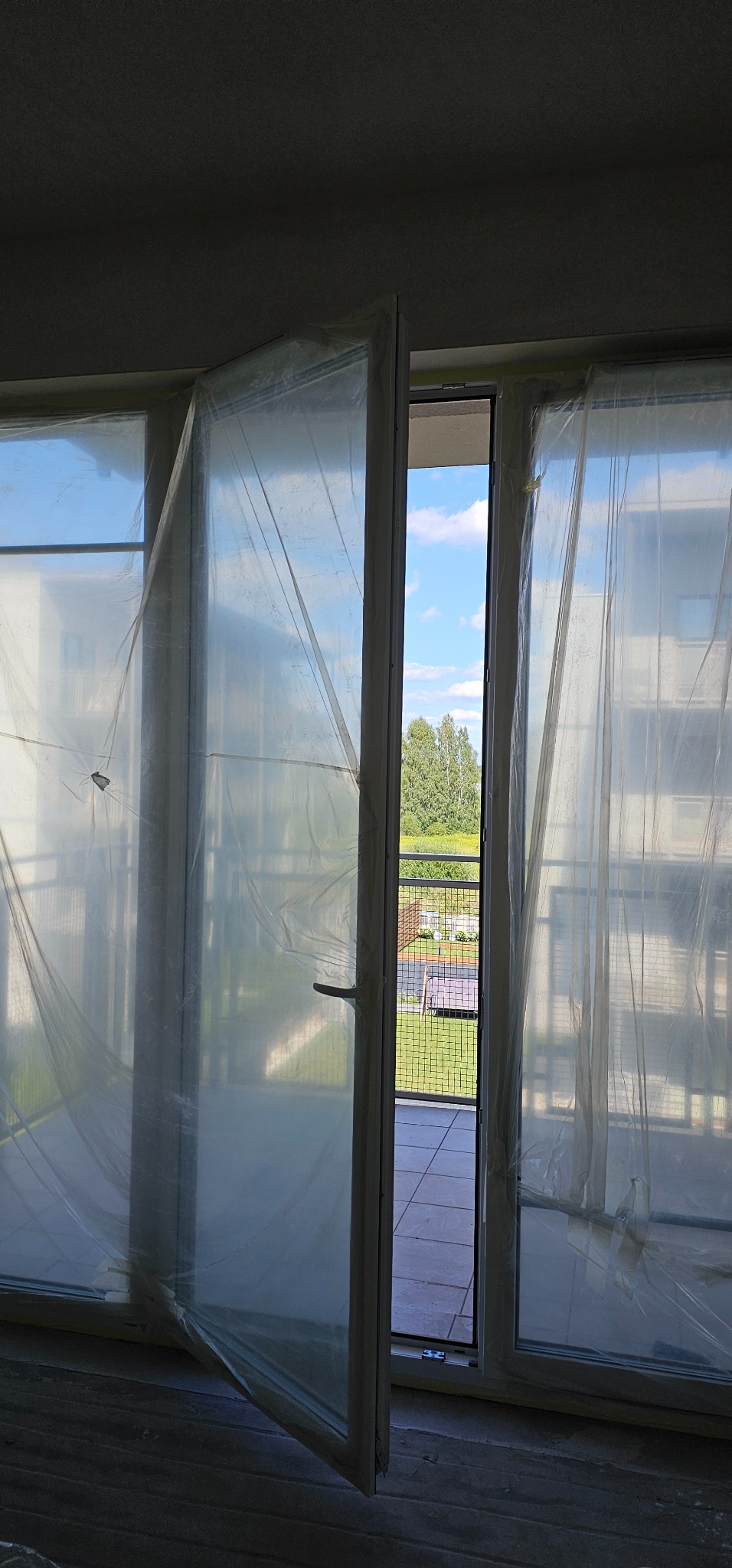 Przygotowanie do prac malarskich: Okna i framugi zabezpieczone folią malarską, otwarte drzwi balkonowe z widokiem na zewnątrz.