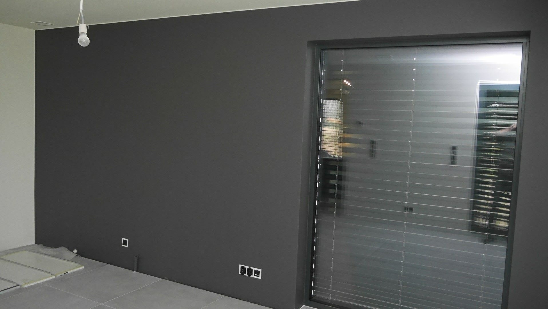 Świeżo pomalowane na szaro wnętrze z widocznymi gniazdkami, fragmentem podłogi z płytkami i oknem z żaluzjami, surowa żarówka zwisa z sufitu.