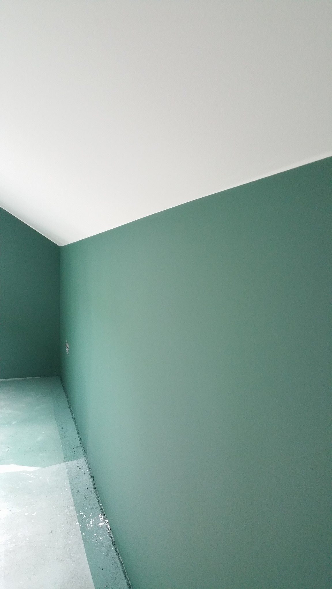 Świeżo pomalowane na zielono ściany i biały sufit w pustym pomieszczeniu, widoczne zabezpieczenia na podłodze przy ścianie.