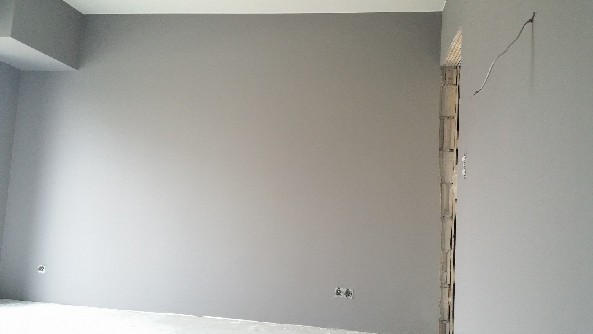 Świeżo pomalowane na szaro wnętrze z gładkimi ścianami, widoczne gniazdka elektryczne i wystające przewody przy futrynie drzwi.