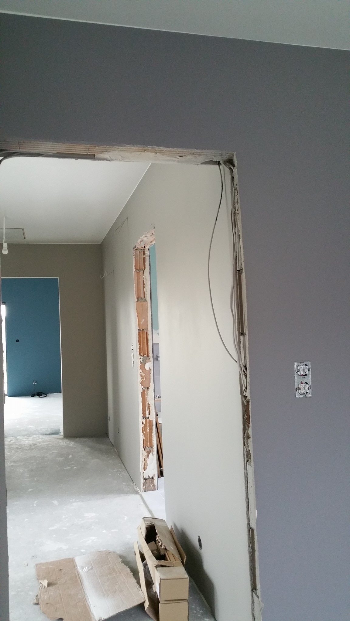 Wnętrze w trakcie remontu: ściany w dwóch odcieniach szarości, fragment ściany z odsłoniętą cegłą po demontażu futryny, widoczne kable elektryczne, podłoga zabezpieczona folią.