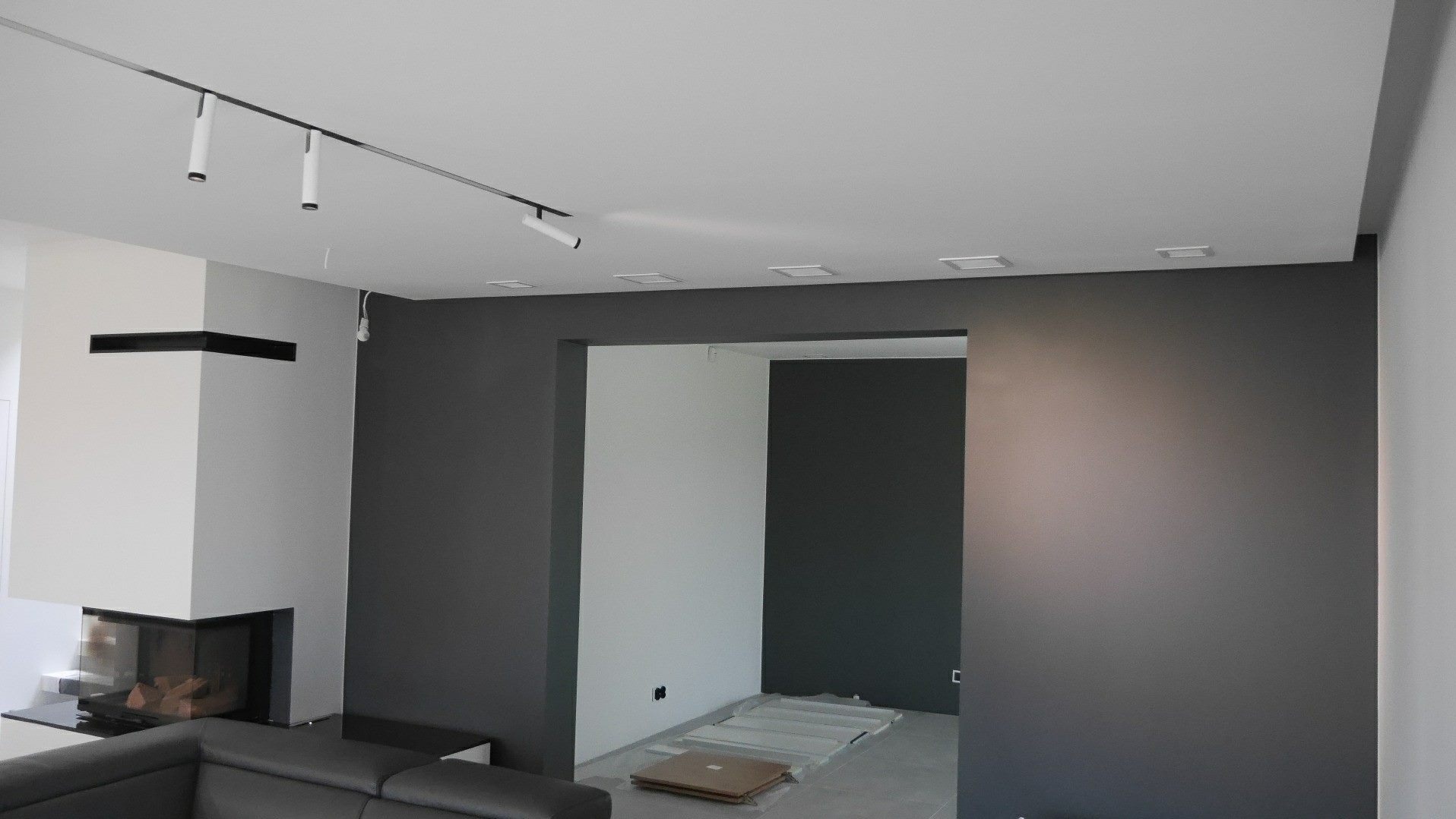 Wnętrze salonu po remoncie z pomalowanymi na szaro ścianami, białym kominkiem z czarnymi akcentami i szynoprzewodem z reflektorami na suficie; widoczne niezabezpieczone gniazdka elektryczne...