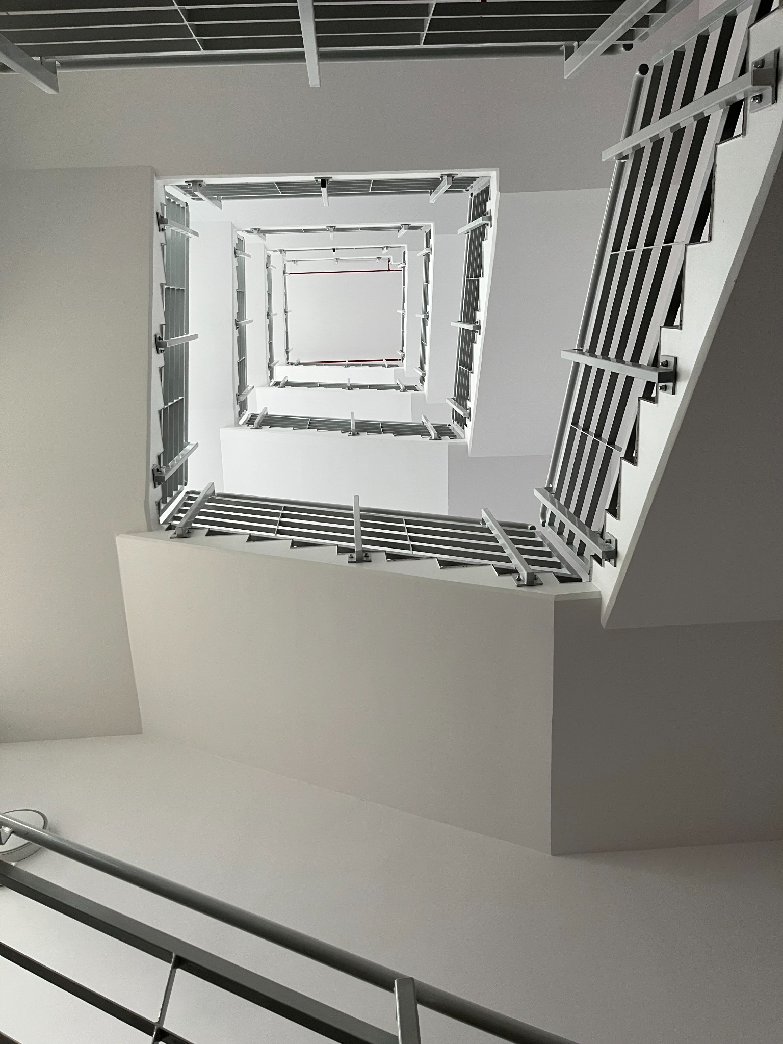 Wnętrze klatki schodowej z widokiem w górę, białe ściany i metalowe barierki, perspektywa z dołu tworzy efekt tunelu.