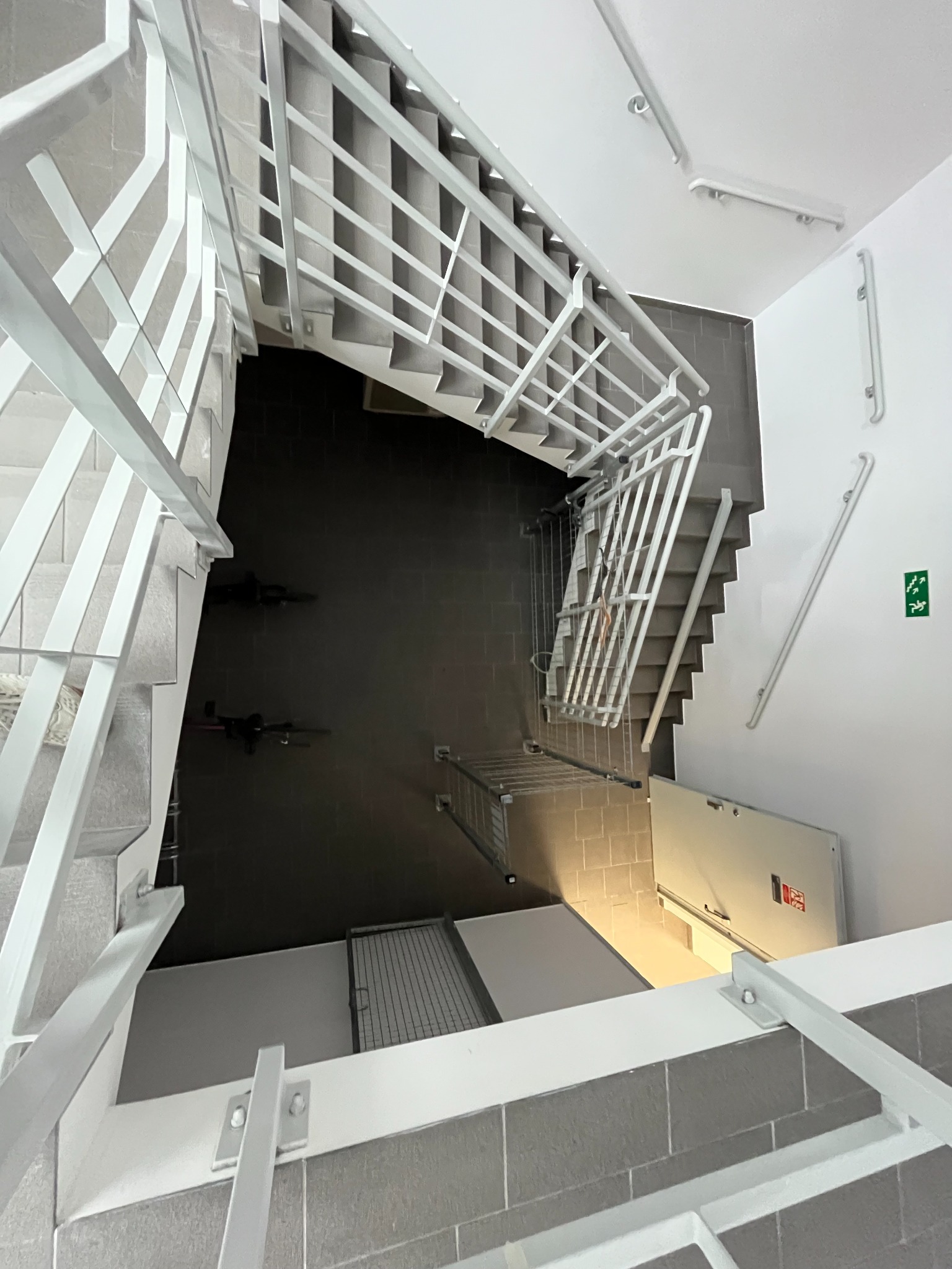 Wnętrze klatki schodowej z widokiem z góry, ukazujące białe metalowe balustrady, szare płytki na podłodze i schodach, oraz awaryjne wyjście po prawej stronie, z dodatkowymi poręczami przy ścianie