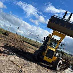 Mfirma Dawid Marach - Żółta ładowarka teleskopowa New Holland z podniesioną łyżką na placu budowy, świeżo usypany nasyp ziemny w tle, widoczne słupy trakcji elektrycznej i błękitne niebo z chmurami.