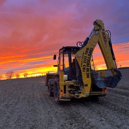 Mfirma Dawid Marach - Żółta koparko-ładowarka New Holland na polu ornym pod dramatycznym, pomarańczowo-fioletowym niebem o zachodzie słońca, widok z tyłu.