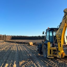 Mfirma Dawid Marach - Żółto-czarna koparko-ładowarka New Holland na polu z przygotowanym terenem pod budowę, w tle widoczny las i czerwony samochód z osobą.