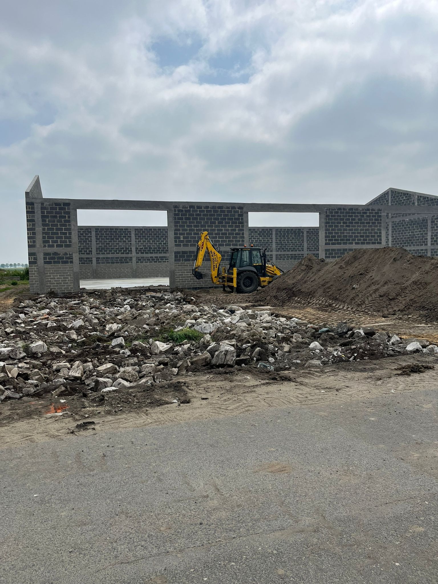 Żółta koparko-ładowarka przy wznoszonej konstrukcji z bloczków betonowych, gruz i ziemia w tle, etap budowy hali.