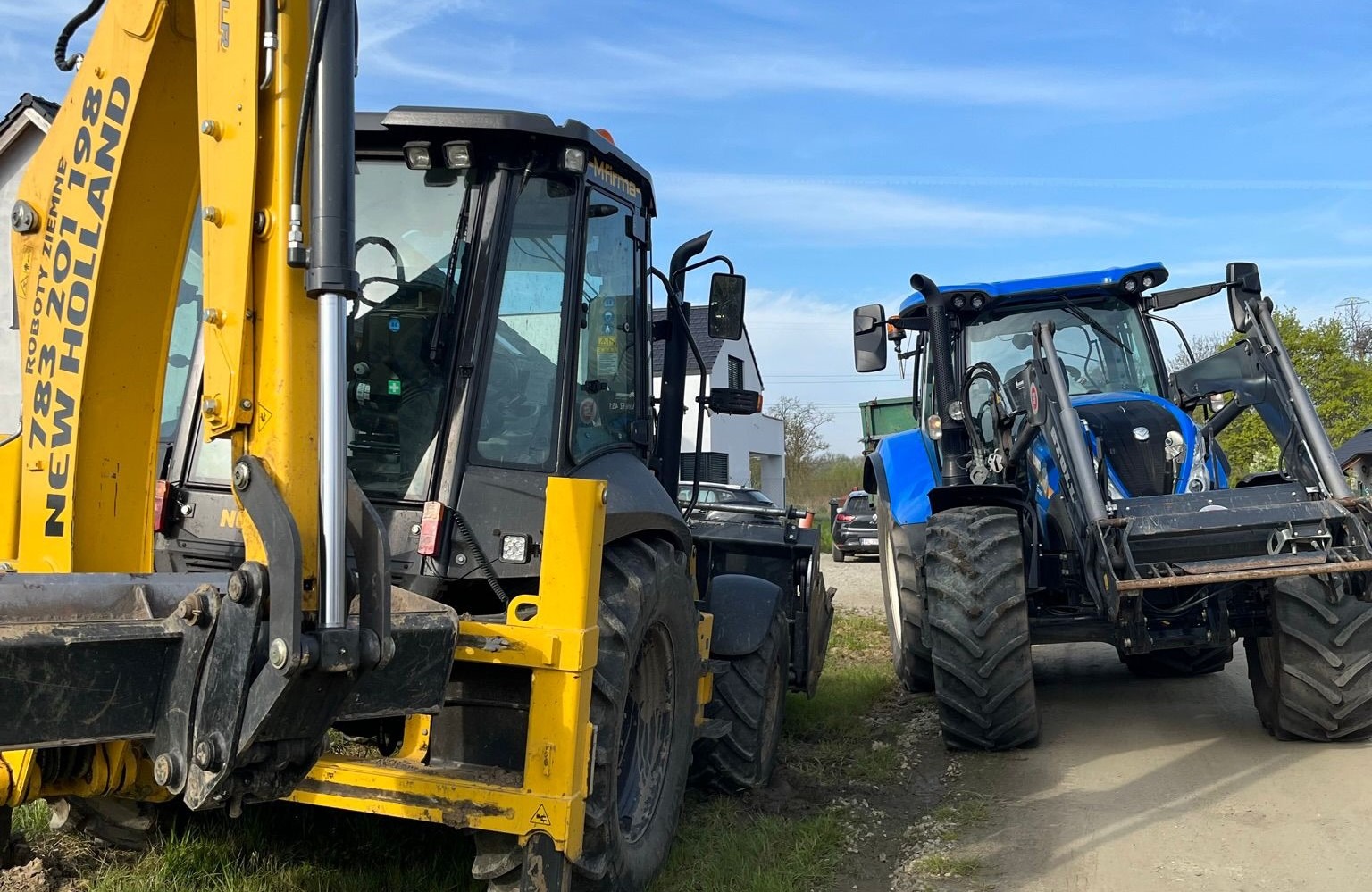 Żółto-czarna koparko-ładowarka New Holland oraz niebieski traktor na tle domu i błękitnego nieba.