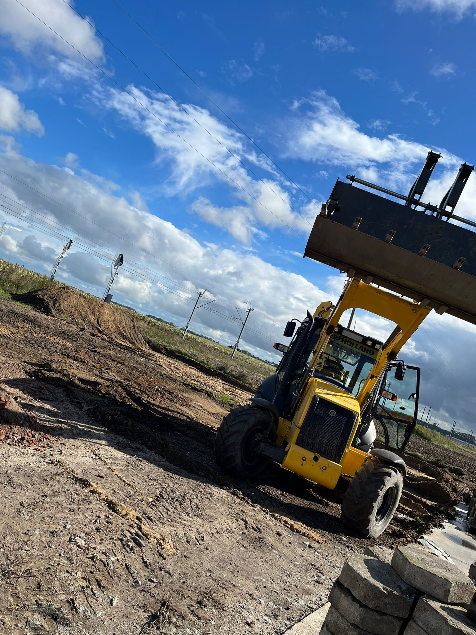 Żółta ładowarka teleskopowa New Holland z podniesioną łyżką na placu budowy, świeżo usypany nasyp ziemny w tle, widoczne słupy trakcji elektrycznej i błękitne niebo z chmurami.