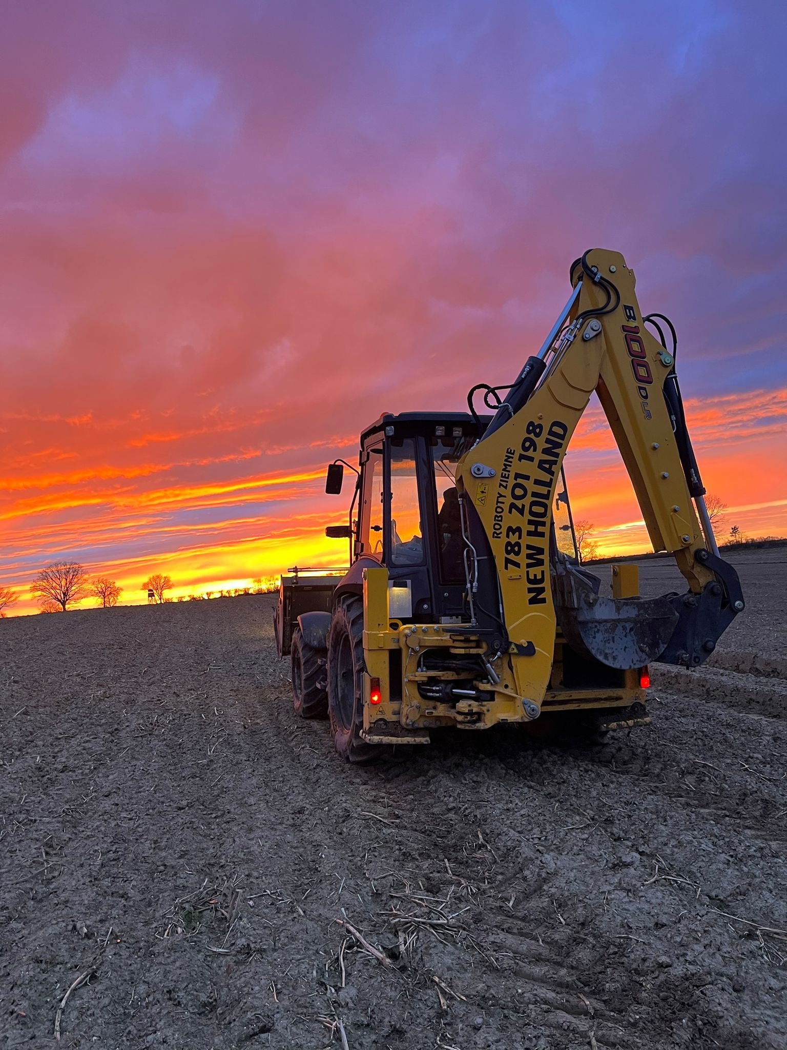 Żółta koparko-ładowarka New Holland na polu ornym pod dramatycznym, pomarańczowo-fioletowym niebem o zachodzie słońca, widok z tyłu.