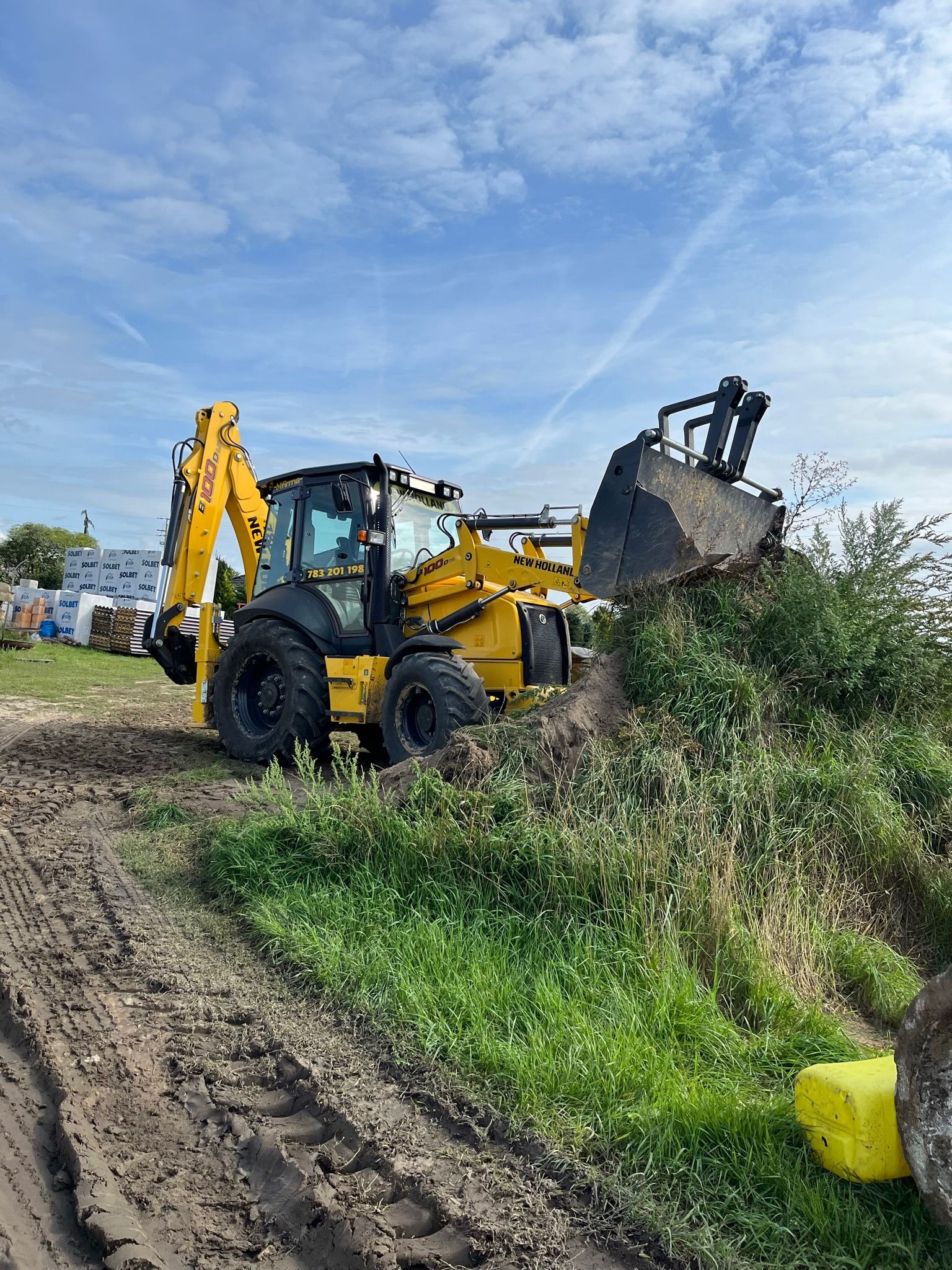 Żółta koparko-ładowarka New Holland nabiera ziemię zarośniętą trawą i krzakami, w tle palety z bloczkami Solbet, błękitne niebo z delikatnymi chmurami.
