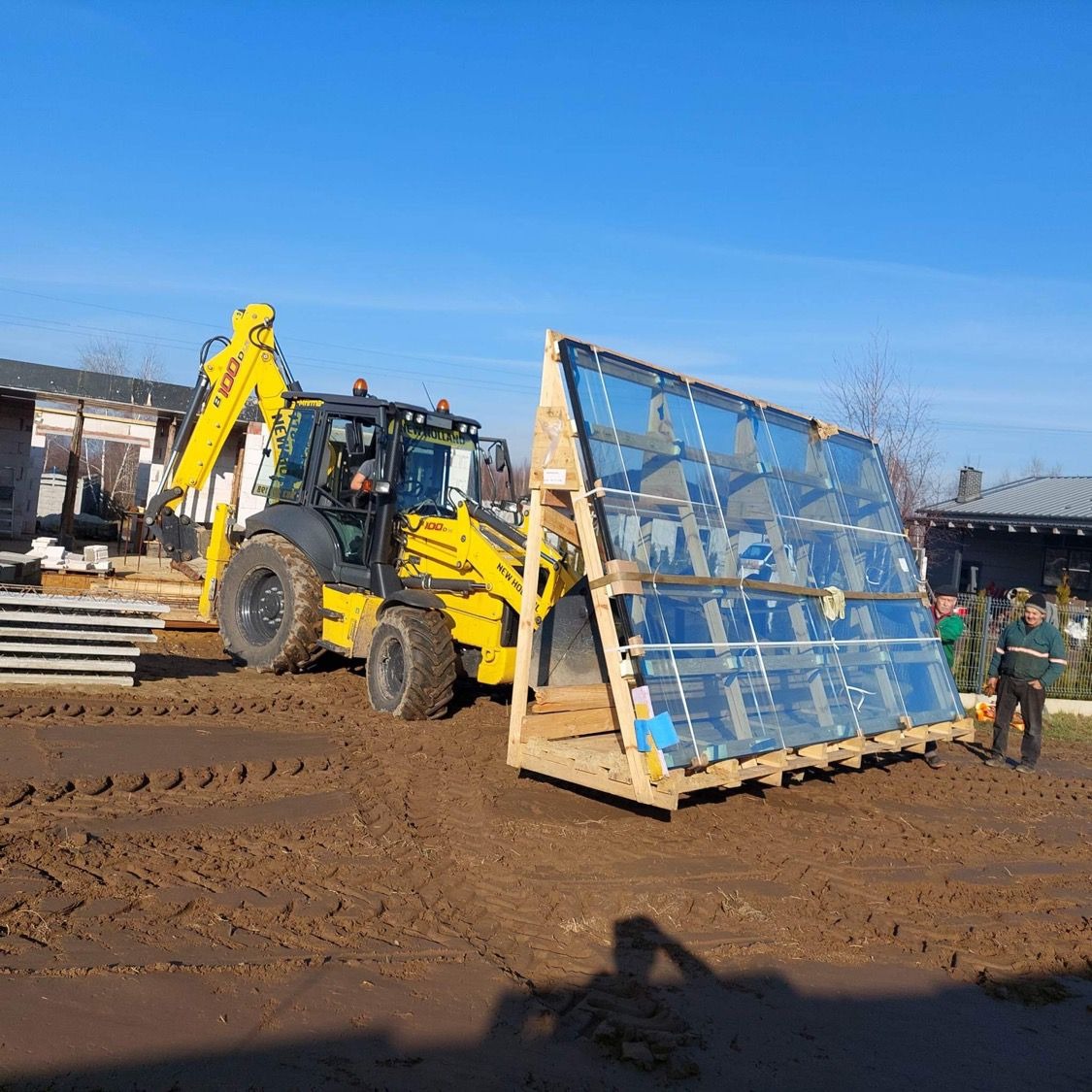 Żółta koparko-ładowarka New Holland transportuje paletę z dużymi taflami szkła budowlanego na placu budowy; dwóch pracowników w tle nadzoruje transport.