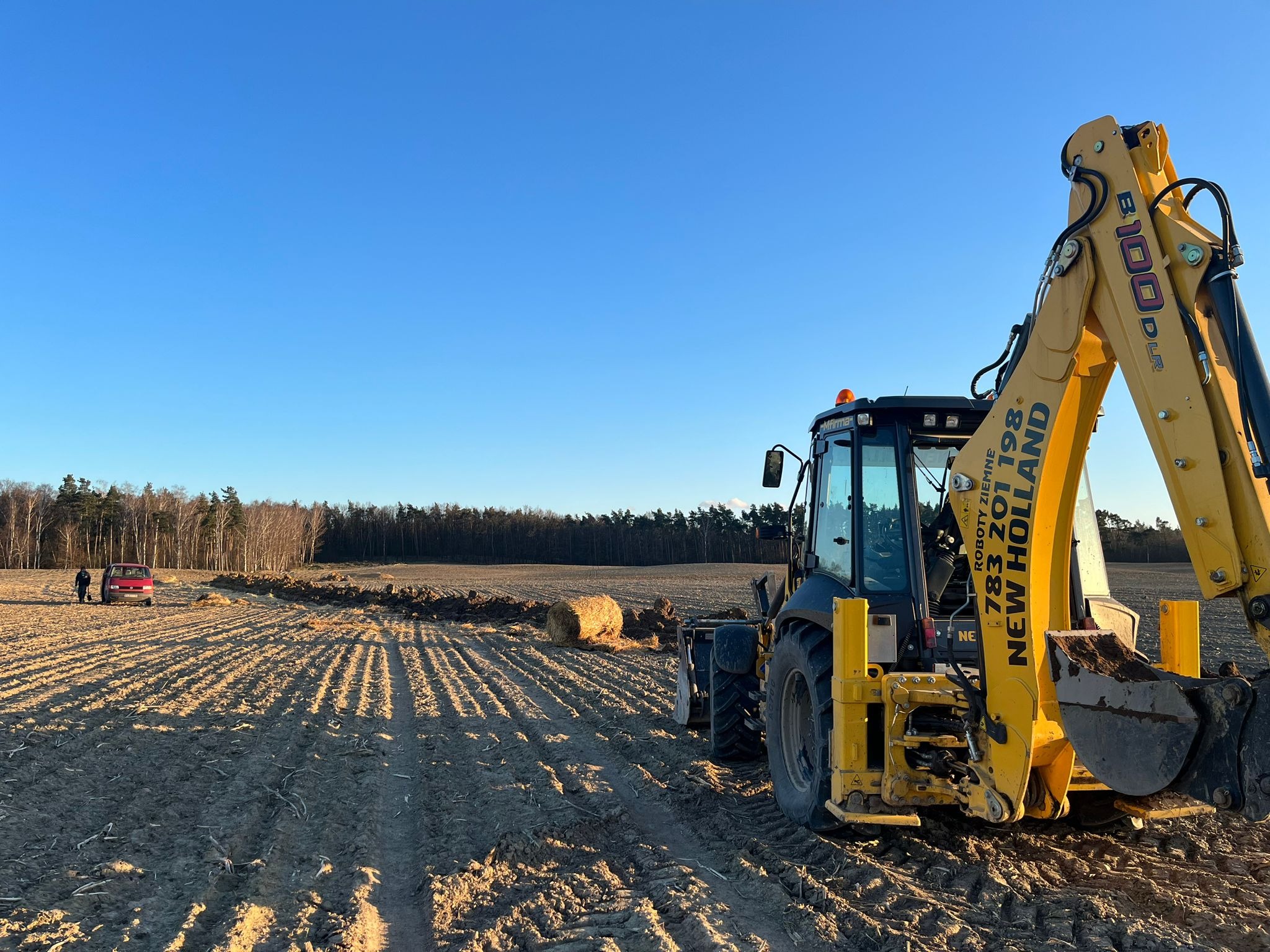 Żółto-czarna koparko-ładowarka New Holland na polu z przygotowanym terenem pod budowę, w tle widoczny las i czerwony samochód z osobą.