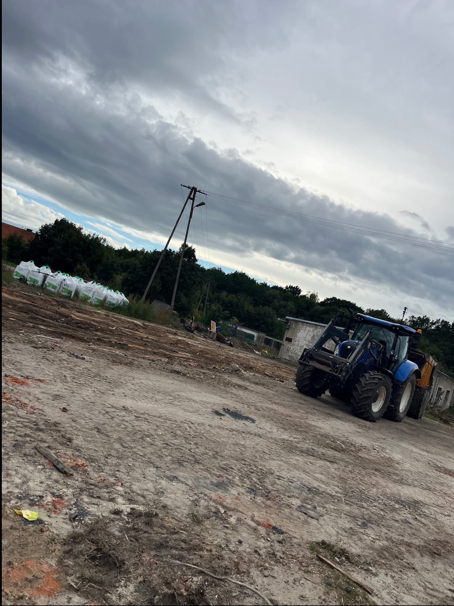 Niebieski traktor z przyczepą na placu budowy, w tle worki z ziemią i słup elektryczny pod pochmurnym niebem. Ujęcie z dołu, pod kątem.