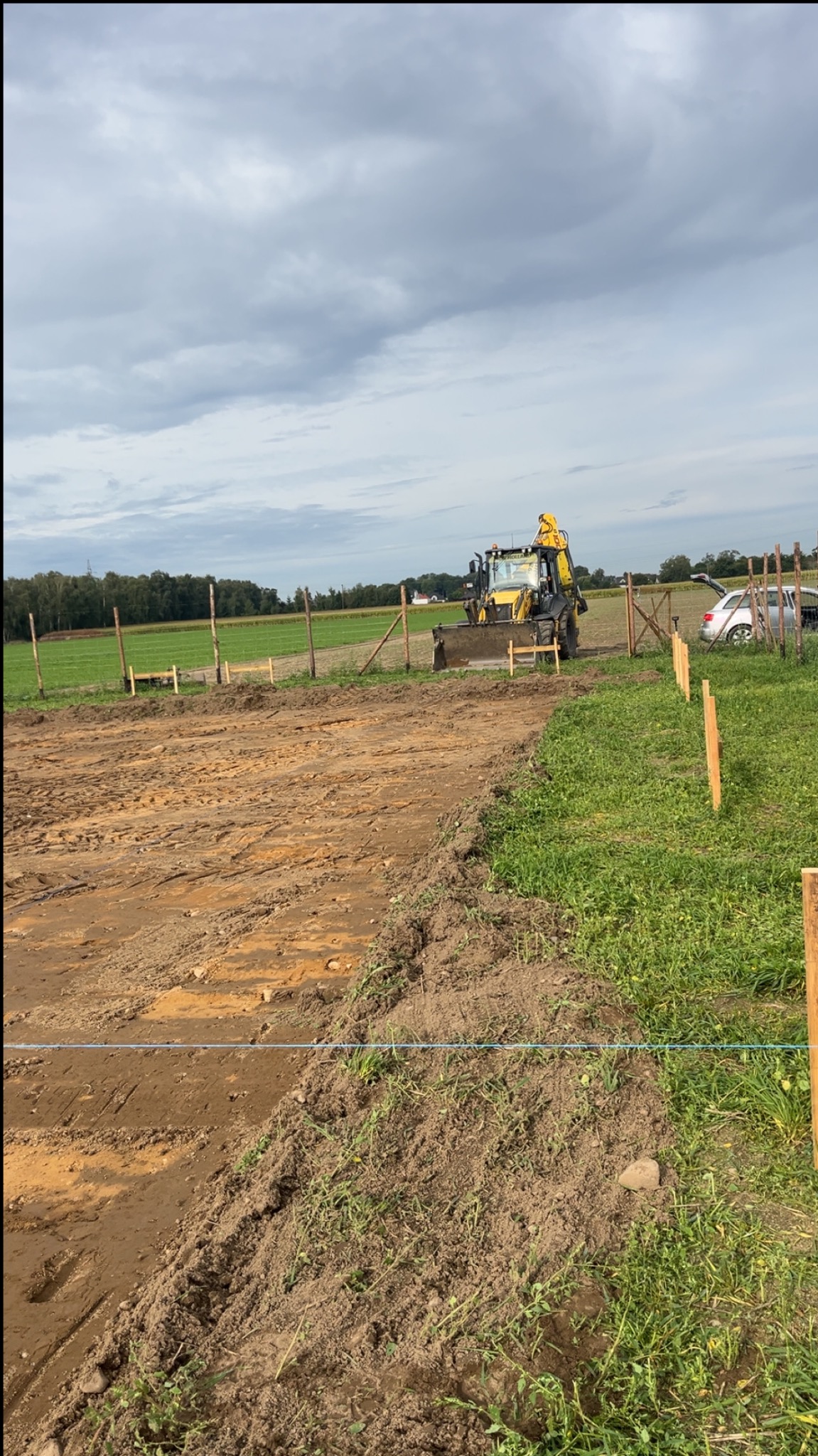 Żółta koparko-ładowarka wyrównuje teren pod budowę, widoczne paliki wyznaczające granice, w tle zielone pola i pochmurne niebo. Prace ziemne w toku.