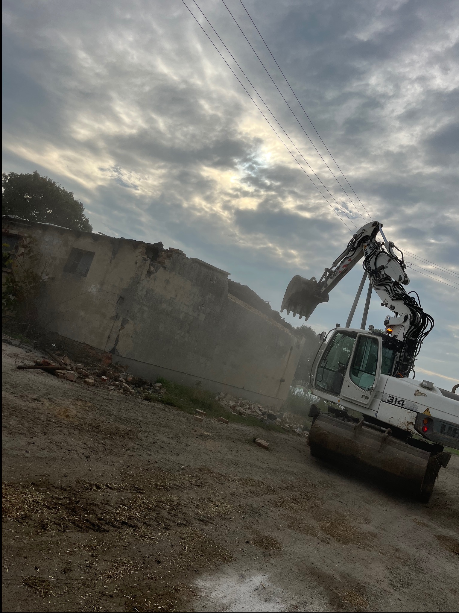 Biała koparka Liebherr 314 w trakcie rozbiórki budynku. Widoczne gruzy i pył, pochmurne niebo. Kąt ujęcia z dołu podkreśla moc maszyny.