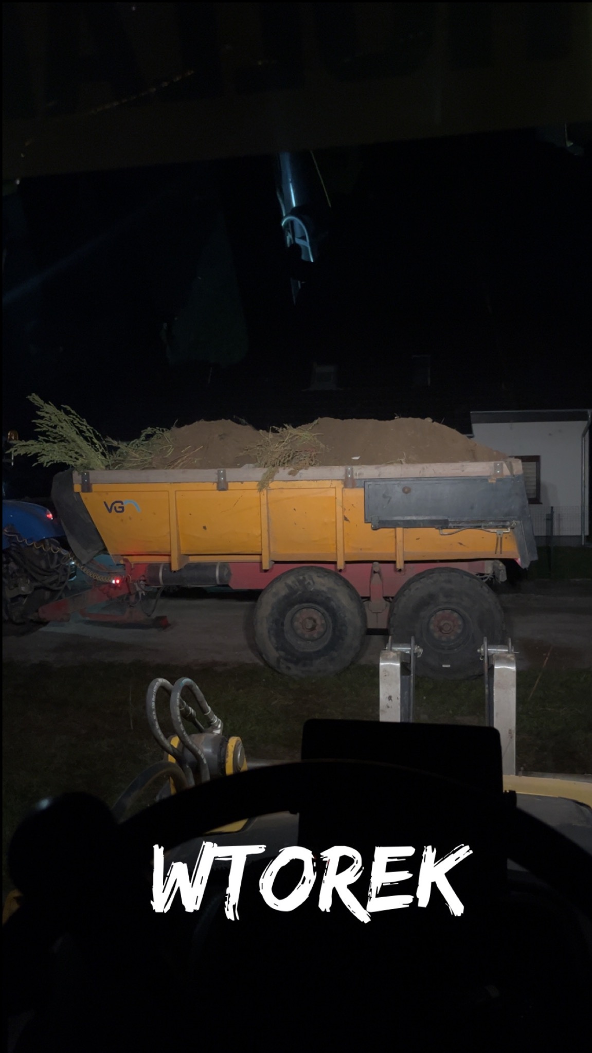 Żółta przyczepa ciągnięta przez traktor, wypełniona ziemią i gałęziami, widziana z perspektywy kierowcy w nocy. Na przyczepie widoczne logo VG.